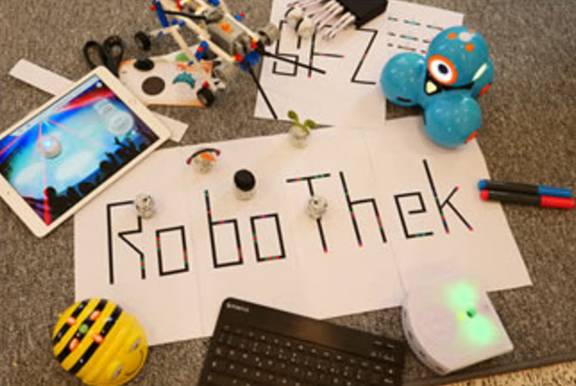 Ein Zettel mit der Schrift Robothek liegt auf einem Tisch mit kleinen MInirobotern daneben.